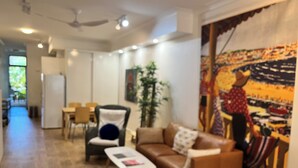 A modern living room with a brown leather sofa, a white coffee table, and a black armchair with a blue cushion. There is a painting on the wall and a ceiling fan above the sofa.
