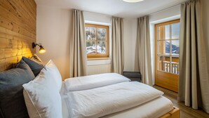 Ein Schlafzimmer mit Holz-Kopfteil, einem Bett mit weißer Bettwäsche, zwei Fenstern mit Vorhängen, einem kleinen Tisch und einer Wandlampe.