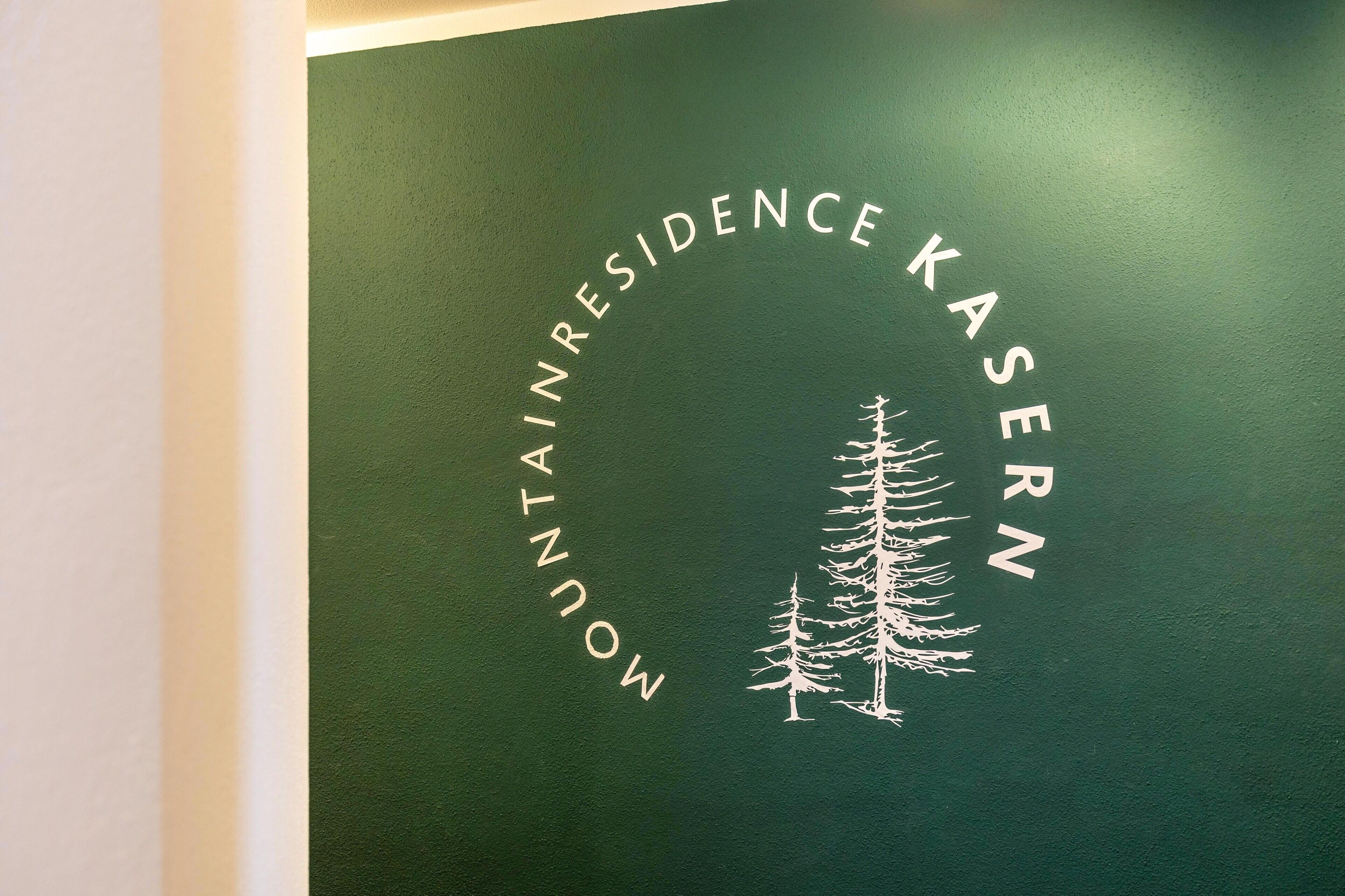A green wall with the text 'MOUNTAIN RESIDENCE KASERN' and a white tree silhouette.