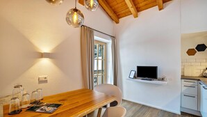 A dining area with a wooden table, chairs, a TV on a shelf, and a window with curtains. - Apartment "Mountain Residence Kasern Nr 4" with Mountain View, Balcony & Wi-Fi (Prettau)