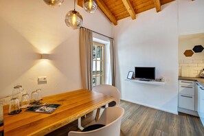 A dining area with a wooden table, chairs, a TV on a shelf, and a window with curtains.