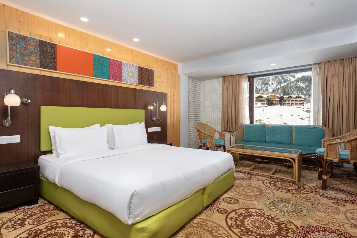 A hotel room with a bed, a sofa, a chair, a small table, and a view of a building and mountains.