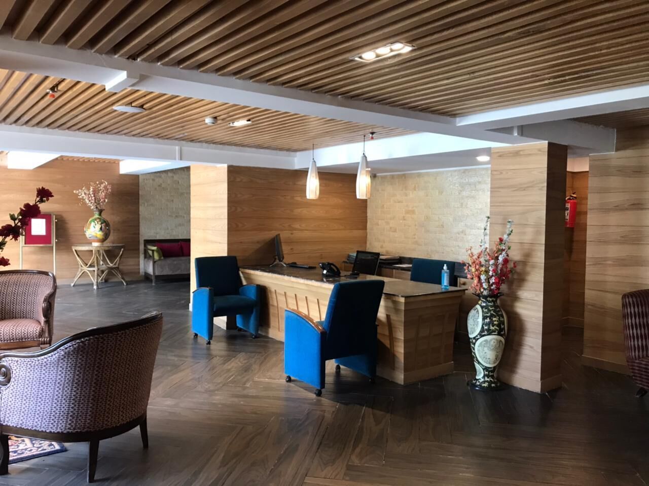 A reception area with a wooden reception desk, blue chairs, a patterned armchair, and a decorative vase with flowers.