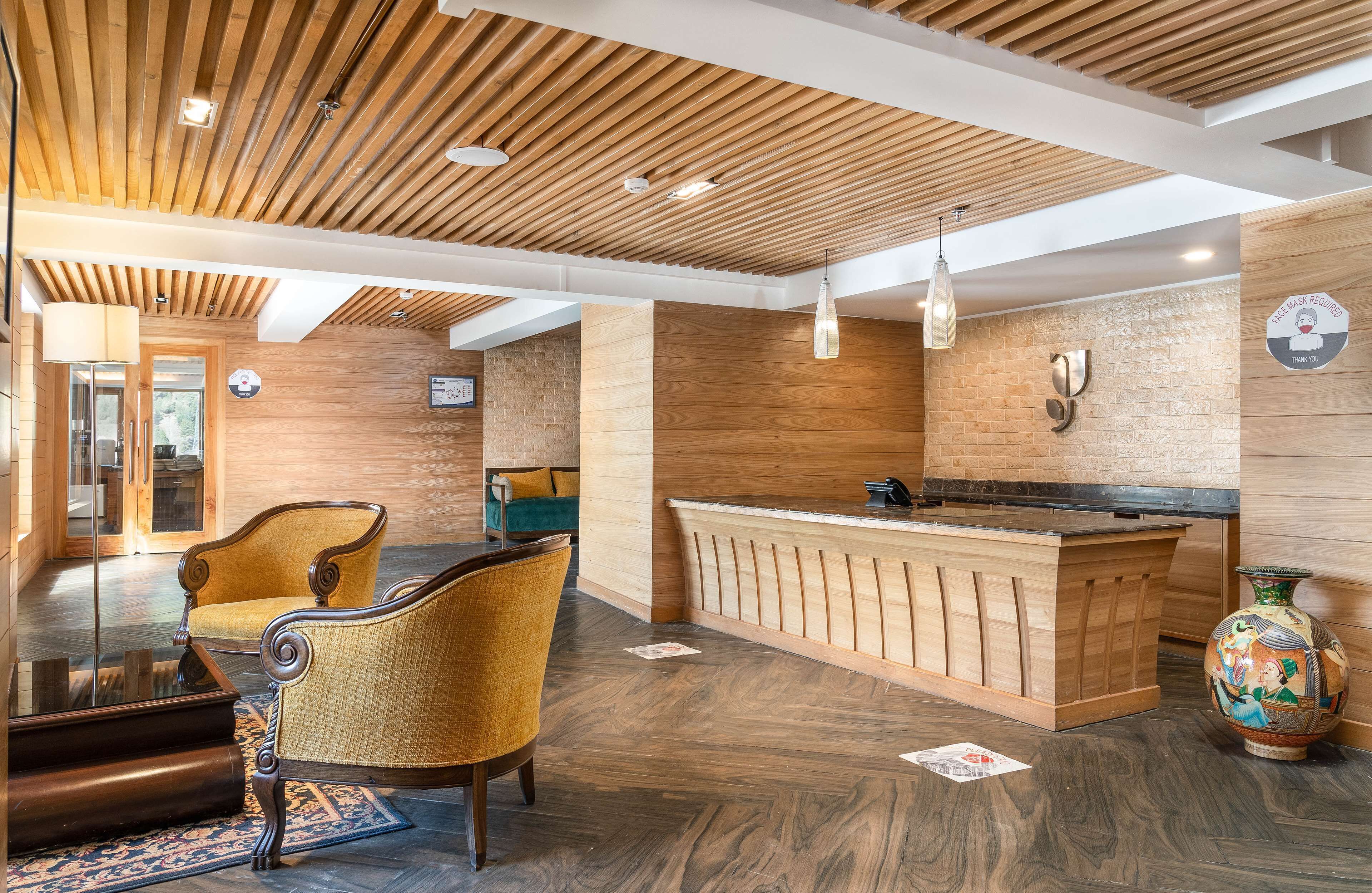 A reception area with a wooden reception desk, two upholstered chairs, a vase, and a wooden ceiling with recessed lighting.