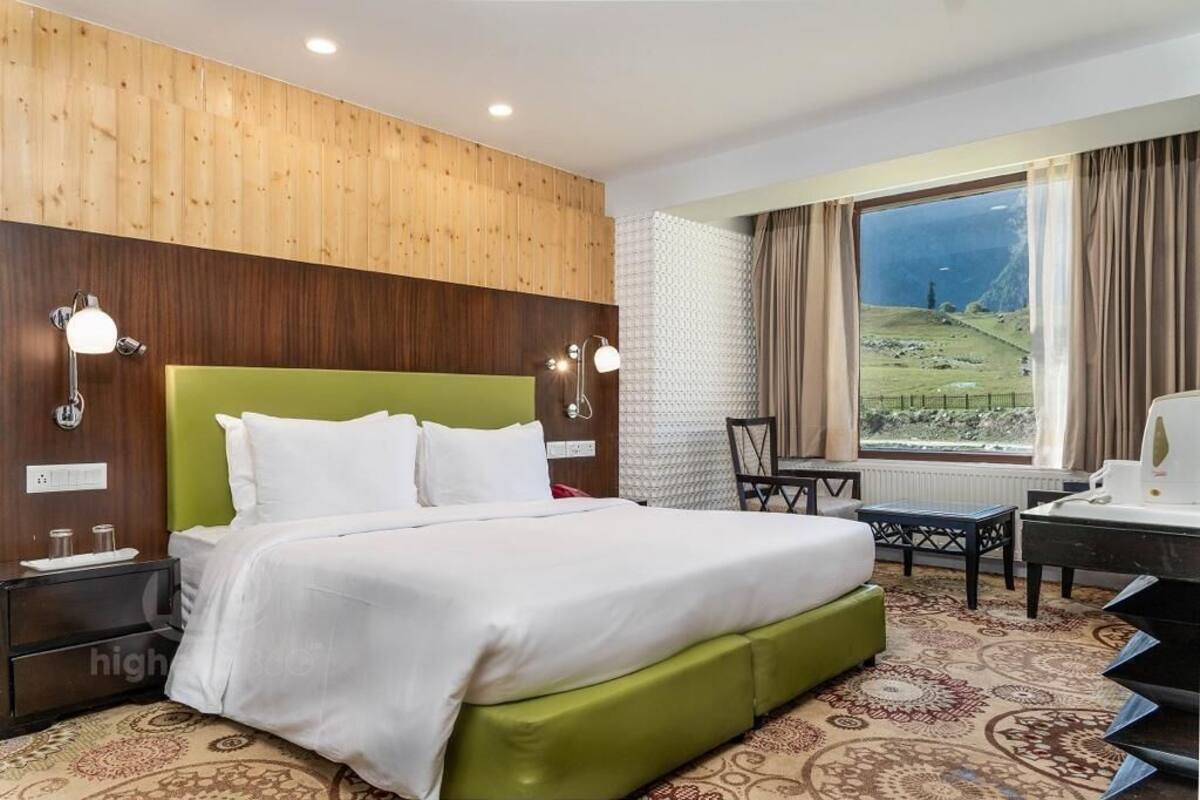 A hotel room with a large bed, a desk, a chair, and a view of a green landscape through the window.