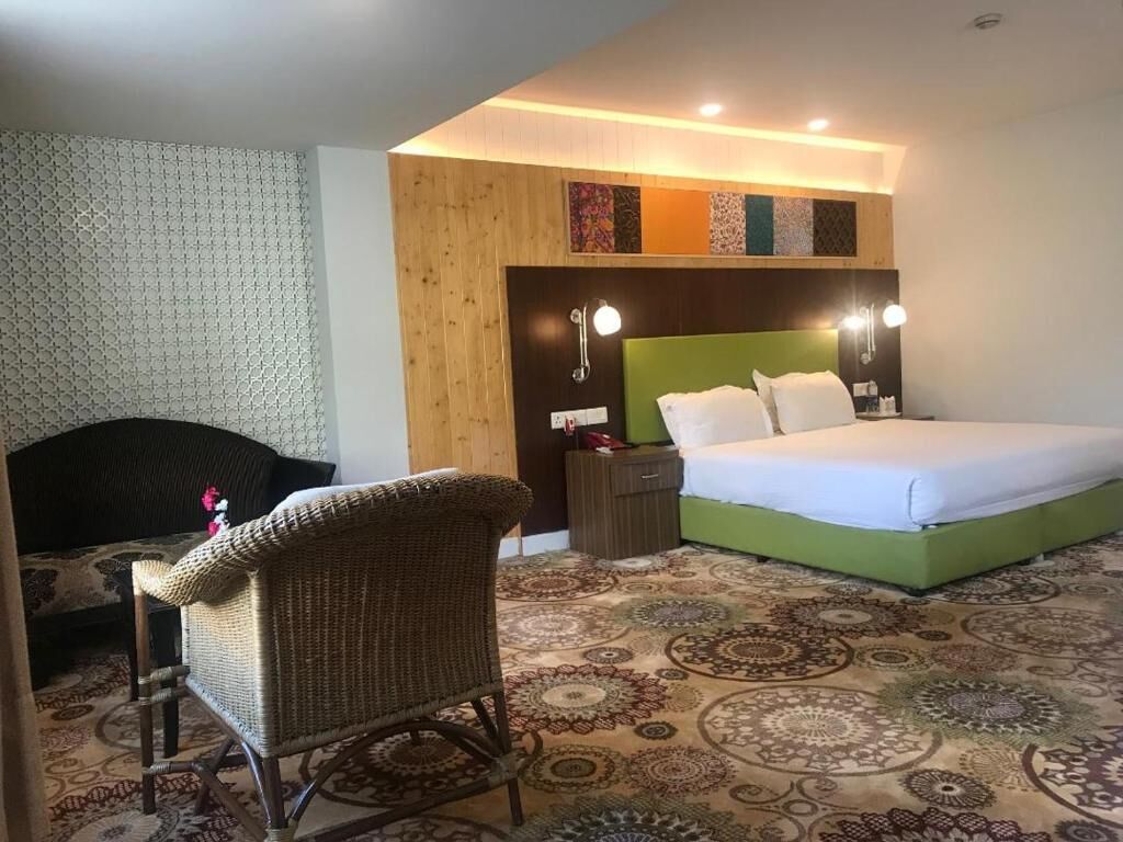 A hotel room with a green bed, a wicker chair, a patterned carpet, and a wooden accent wall.