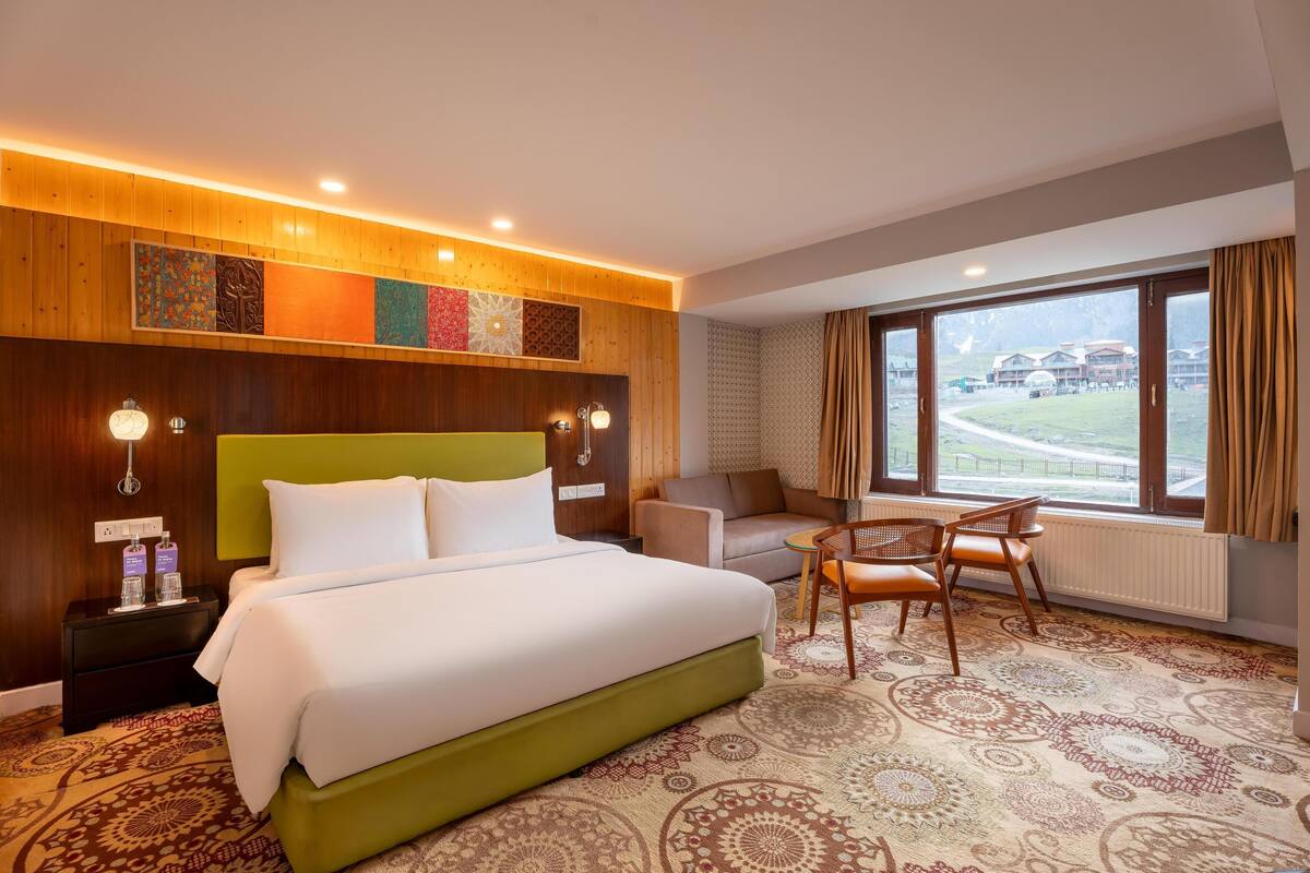A hotel room with a large bed, a sofa, a chair, a window with a view, and a patterned carpet.