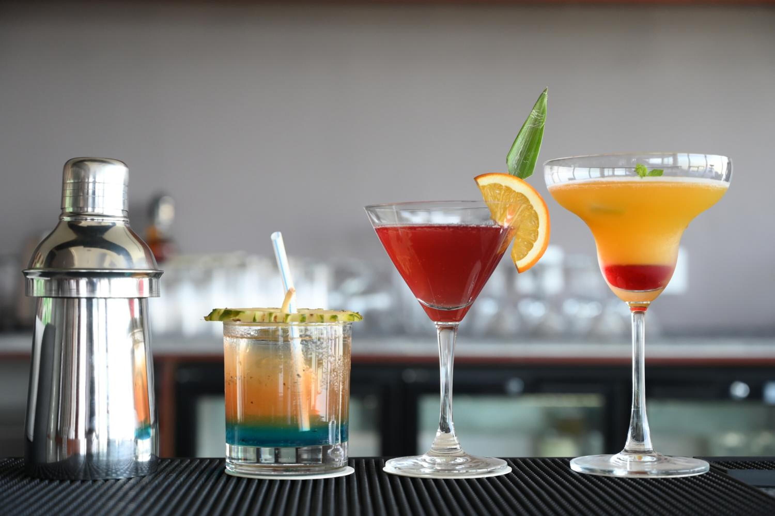 Trois cocktails sont posés sur le comptoir d’un bar, accompagnés d’un shaker à cocktails et d’un verre décoré d’une tranche de citron vert.
