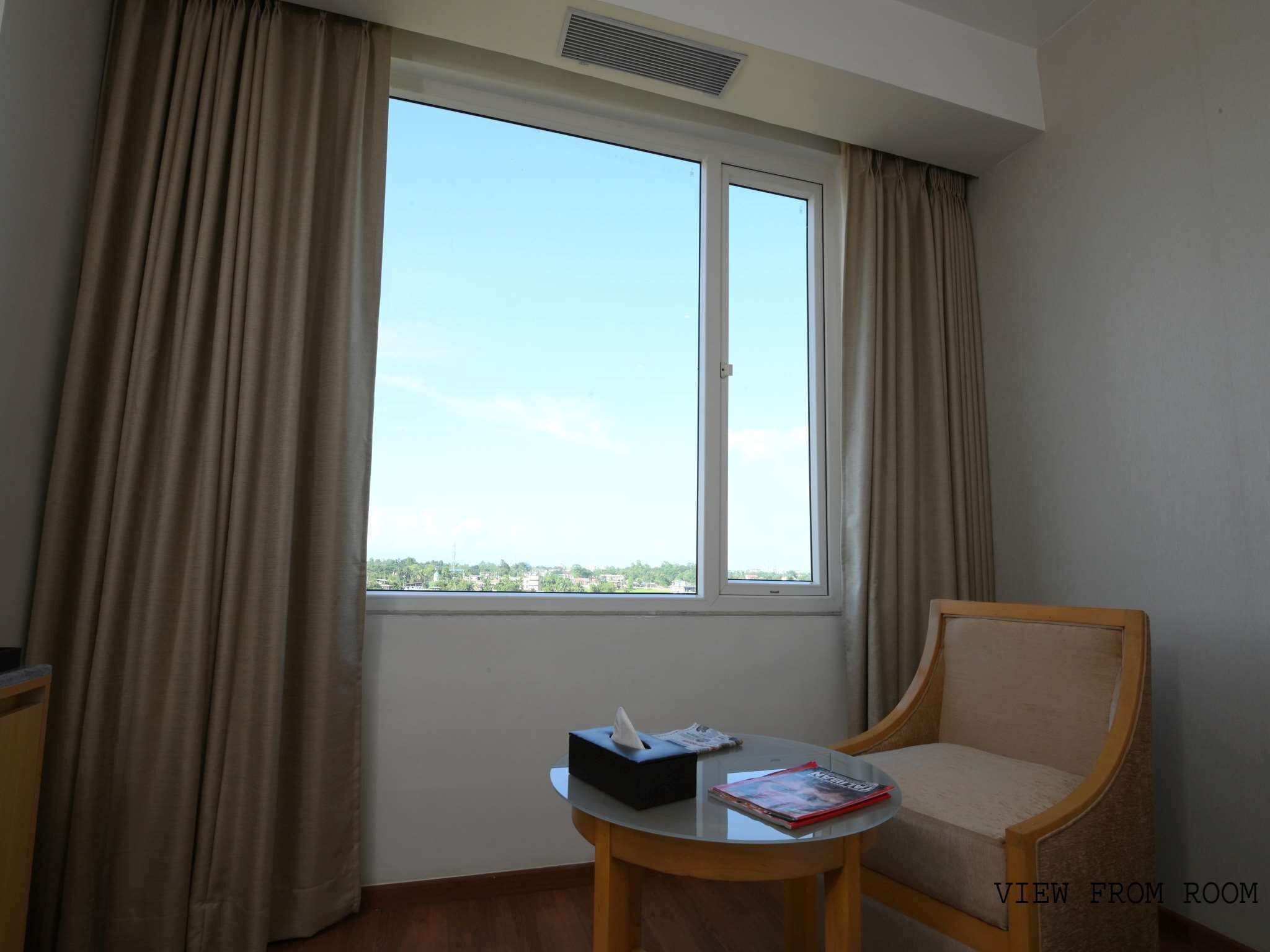 A hotel room with a large window, a chair, and a small table with a tissue box and a magazine.