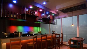 A bar area with wooden stools and a table, shelves with bottles, and a view of the city through large windows.