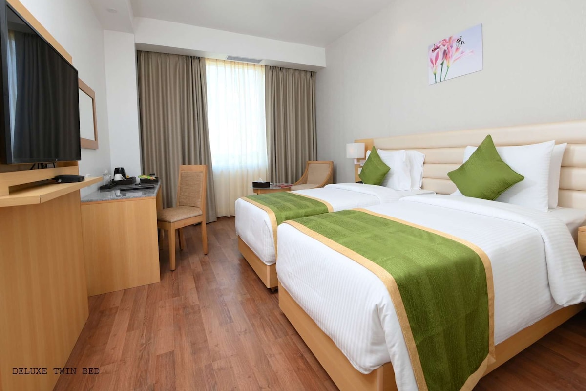 Habitación de hotel con dos camas individuales, un escritorio, una silla, un televisor y una ventana con cortinas.
