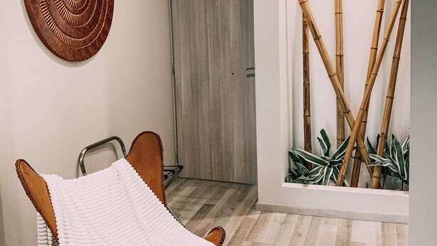 A modern room with a wooden floor, a white chair with a white cover, a wooden door, and a wall with a circular wooden artwork and bamboo plants.