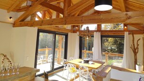 Un intérieur en bois avec un plafond haut, de grandes fenêtres et un coin repas avec des chaises blanches et une table.