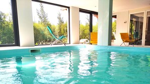 Une piscine intérieure à l’eau claire, dotée de grandes fenêtres et offrant une vue sur la verdure environnante.