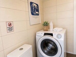 A hotel bathroom with a washing machine, a toilet, a 'Do Not Throw Trash or Paper Towels in Toilet' sign, and a framed artwork on the wall.