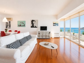 A modern living room with a white sofa, a coffee table, and a TV. There are red chairs, a dining table, and artwork on the walls. A balcony with a view of the sea is visible. - Adelos Coastal Chic by the Sea - Athens Riviera (Alimos)