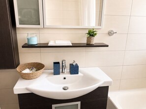A bathroom with a white sink, a mirror, a shelf with a plant, and a basket.