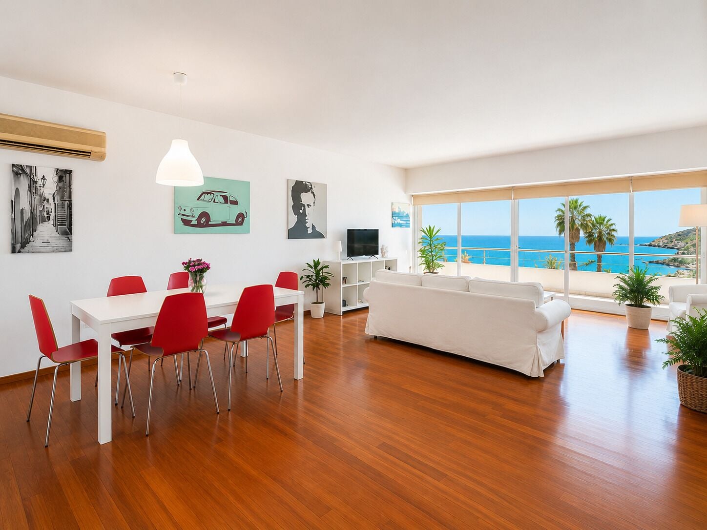 Una sala de estar moderna con un sofá blanco, sillas rojas, una mesa de comedor y un amplio ventanal con vista al mar.