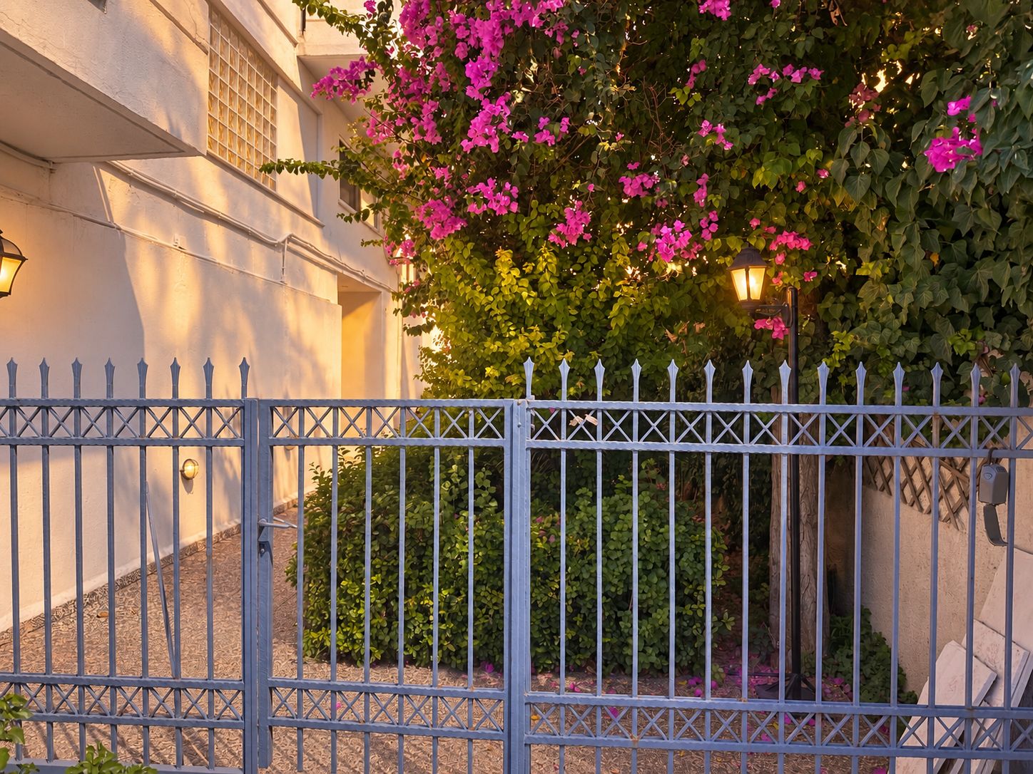 A gated entrance with a metal fence, a building with a light-colored facade, and a flowering bush.