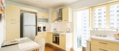 A modern kitchen with a refrigerator, oven, and coffee maker. There is a dining table with chairs and a window with a view of buildings.