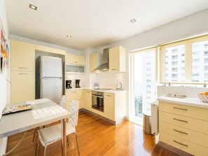 Uma cozinha moderna com geladeira, forno e cafeteira. Há uma mesa de jantar com cadeiras e uma janela com vista para edifícios.