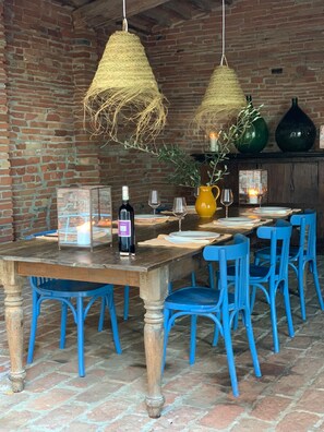 Un coin repas rustique avec un mur en briques, une table en bois, des chaises bleues, des luminaires suspendus et un vase agrémenté de verdure.