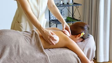 A person receiving a back massage in a room with a bed covered in a beige towel, a small table with a plant, and a curtain in the background.