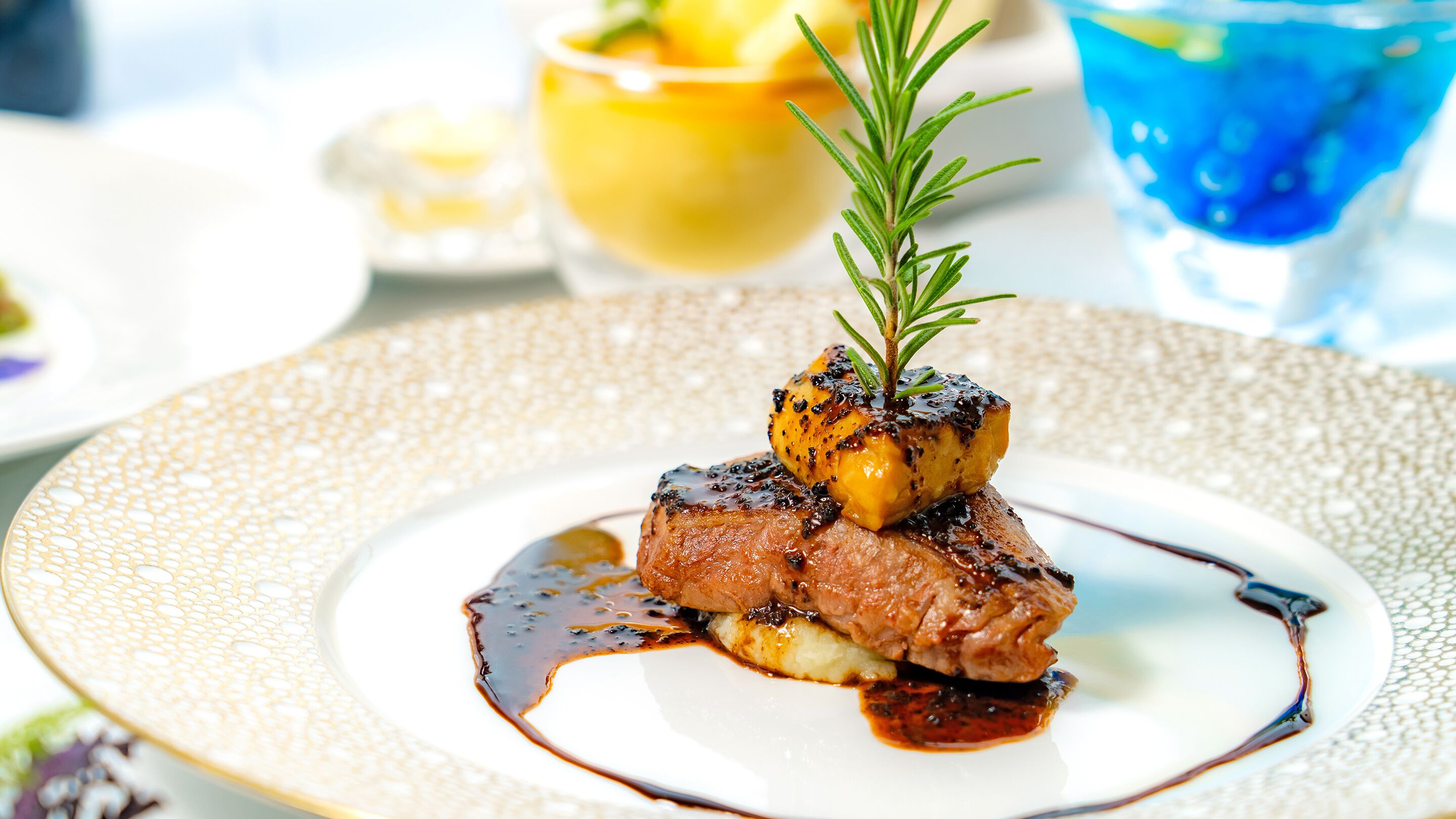 Un plat composé de viande et de légumes, décoré d’une branche de romarin, présenté sur une assiette blanche à motifs.
