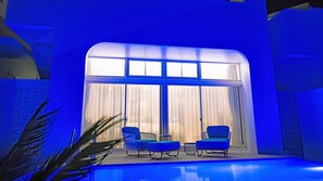 A modern indoor pool area with blue lighting, two lounge chairs, and a large window with curtains. - Blue Ocean Hotel & Resort Miyakojima (Miyakojima)