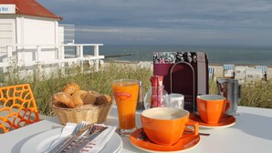 Ein Frühstückstisch mit orangenen Tassen und einem Brotkorb, mit Blick auf einen Strand, im Hintergrund ein weißes Gebäude.