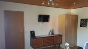 Una habitación con un armario de madera, un televisor montado en la pared, una mesa de comedor con sillas y una planta en maceta sobre la mesa.