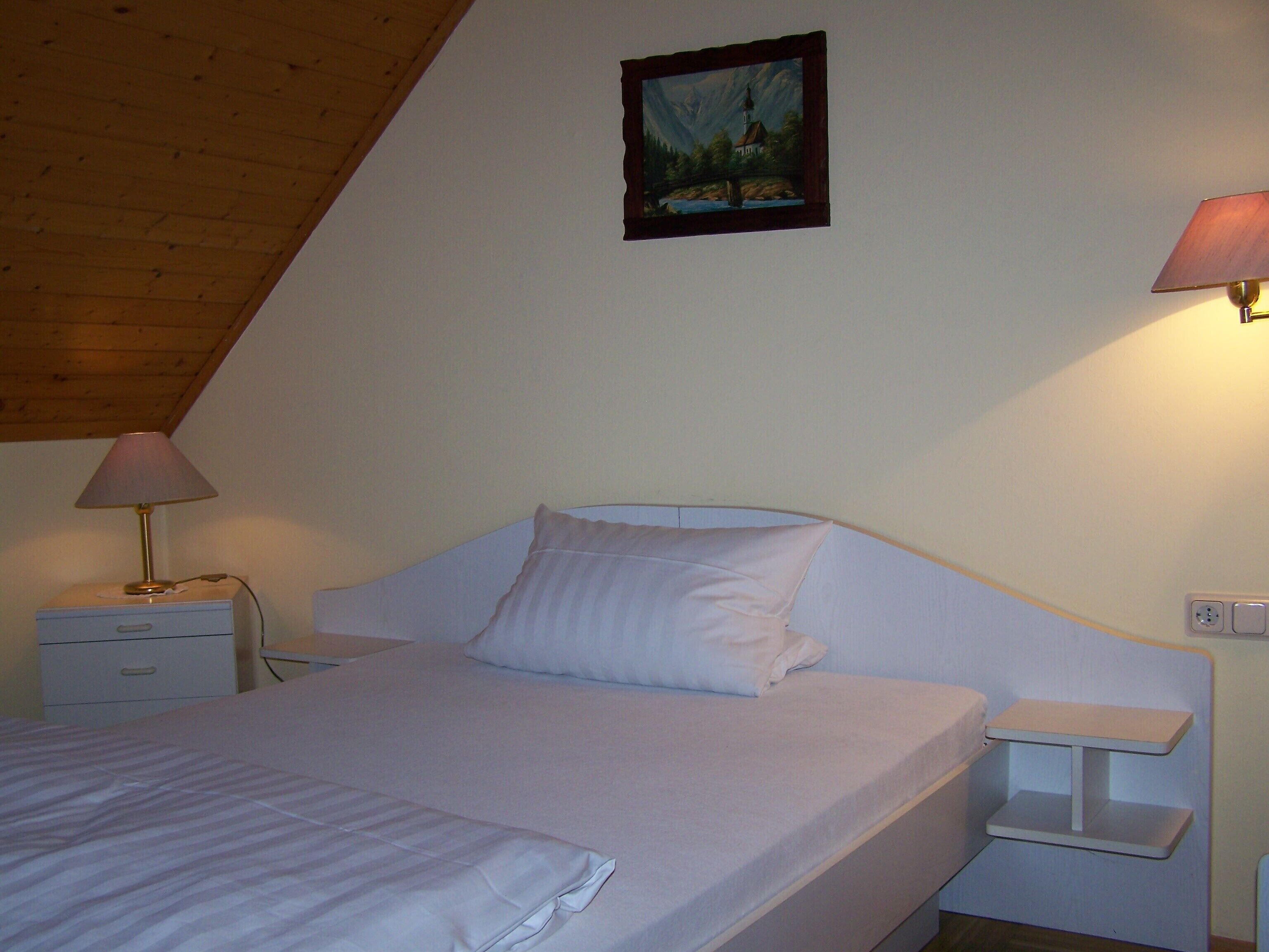 Een slaapkamer met een bed, nachtkastje, lamp en een schilderij aan de muur.