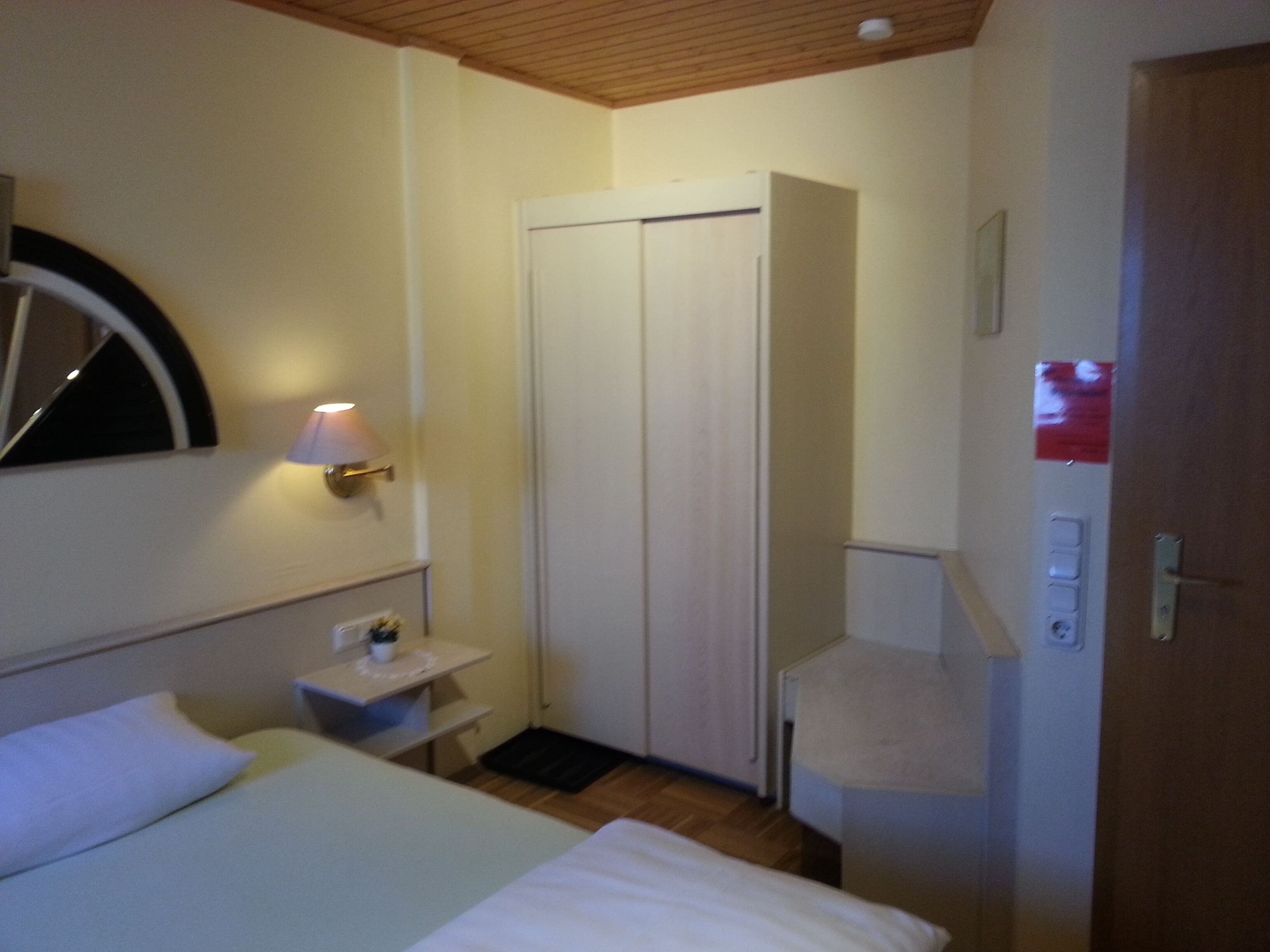 Ein Hotelzimmer mit Bett, Nachttisch, Lampe, Kleiderschrank und einer Tür mit Lichtschalter.
