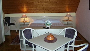 Una habitación pequeña y acogedora con techo de madera, una cama con sábanas blancas, dos lámparas de noche, una mesita con un objeto decorativo y dos sillas.