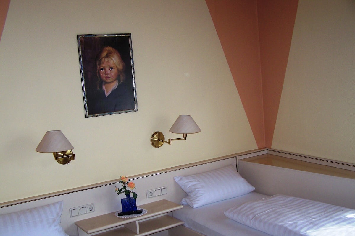 A hotel room with two single beds, a small bedside table with a vase of flowers, two wall-mounted lamps, and a framed picture on the wall.