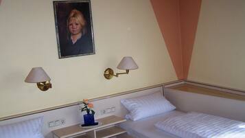A hotel room with two single beds, a small bedside table with a vase of flowers, two wall-mounted lamps, and a framed picture on the wall.