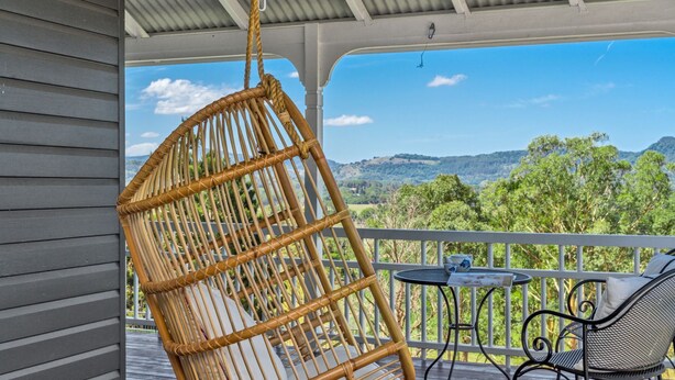 A wicker chair hanging from a ceiling, a metal table, and a view of a green landscape with trees and hills. - The Hideout House - hinterland views for miles! (Myocum)