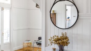 A room with white paneled walls, a round mirror, a wicker stool, a chair, and a vase with dried flowers. - The Hideout House - hinterland views for miles! (Myocum)