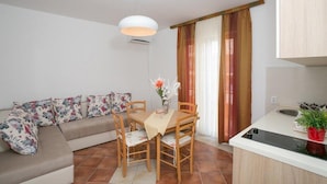 A compact living space with a sofa, dining table, chairs, a pendant light, a kitchen area with a sink, and a potted plant. - Milans Apartments (Pržno)