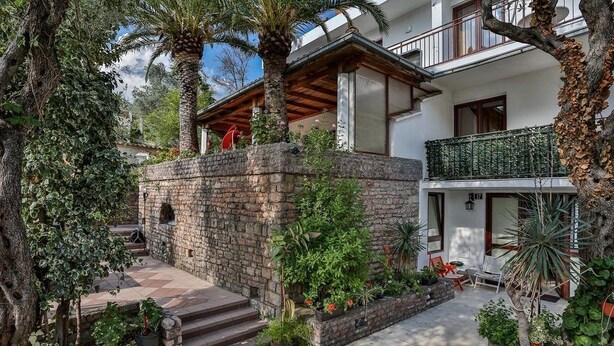 A courtyard with stone steps leading to a wooden deck, surrounded by lush greenery and palm trees. - Milans Apartments (Pržno)