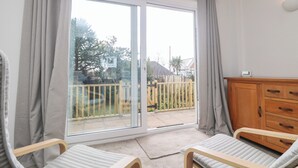 Une pièce comprenant deux chaises en bois, un meuble en bois et une porte-fenêtre coulissante donnant sur un balcon avec vue sur un jardin et des maisons.