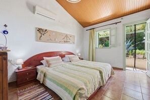 Ein Schlafzimmer mit Holzboden, einem Bett mit gestreifter Bettwäsche, einem Nachttisch mit Lampe, einer Wand-Klimaanlage und einem Fenster mit Vorhängen.