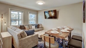 A cozy living room with a sofa, a dining table set with plates and glasses, a TV mounted on the wall, and large windows with a view of the outside. - A Ridge above the Rest!  New Remodel and High End Upgrades (Breckenridge)