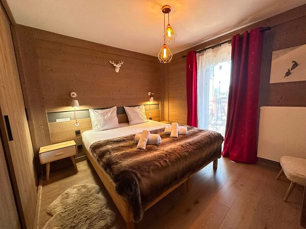 A bedroom with a wooden headboard, a bed with a brown fur throw, a deer head decoration, and a window with red curtains.