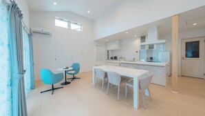 A modern kitchen with white cabinets and a central island, a dining area with a white table and blue chairs, and a large window providing natural light. - Villa Capri Miyakojima (Miyakojima)