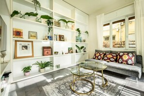 Un salon comprenant un canapé, deux tables basses avec un plateau en verre, un tapis à motifs et une étagère blanche où sont disposées des plantes et des objets décoratifs encadrés.
