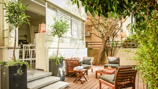 An outdoor patio with wooden furniture, potted plants, and a view of a building with large windows. - Serene Retreat with Lush Garden by Sea N Rent (Tel Aviv)