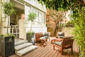 Un patio al aire libre con mobiliario de madera, plantas en macetas y vistas a un edificio con amplias ventanas.