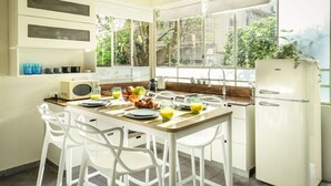 A kitchen with a dining table set for breakfast, white chairs, a refrigerator, and a window with a view of greenery. - Serene Retreat with Lush Garden by Sea N Rent (Tel Aviv)