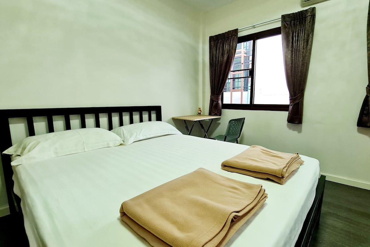 A neatly made bed with white linens and two folded brown towels, a small table with a chair, and a window with curtains.